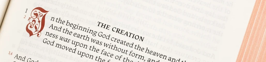 A close view of the open Reader&rsquo;s Edition at the first verses of Genesis: a red decorated initial, the heading THE CREATION in small capitals, the opening lines running as a single column of prose with the verse numbers small in the margin, and the art-gilt edges showing pink down the right.